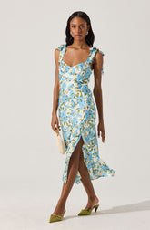 The woman is wearing a sleeveless, ankle-length dress with a light-colored background and a floral pattern featuring blue, white, and green colors. The dress has shoulder straps tied into bows. She is also wearing green pointed-toe high heels and carrying a small, white handbag.